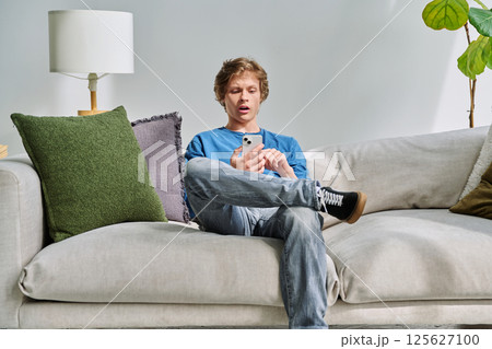 Handsome surprised teenage guy using smartphone sitting on couch at home Handsome surprised teenage guy using smartphone sitting on couch at home 125627100