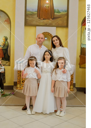 A festive family has gathered in the church at the moment of an important event for the eldest child. Two twin girls and their sister in elegant attire stand beside their mother and father. A festive family has gathered in the church at the moment of an important event for the eldest child. Two twin girls and their sister in elegant attire stand beside their mother and father. 125627468