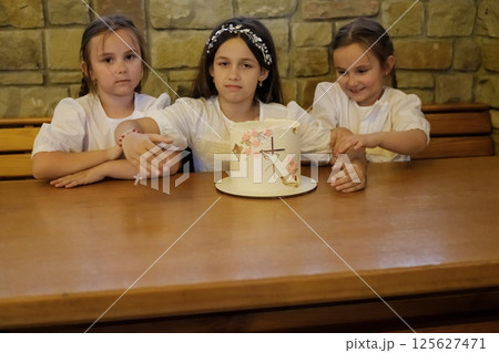 Little girls sit at the table, admiring the festive cake. The sisters look joyful and full of anticipation for the sweet treat. Little girls sit at the table, admiring the festive cake. The sisters look joyful and full of anticipation for the sweet treat. 125627471