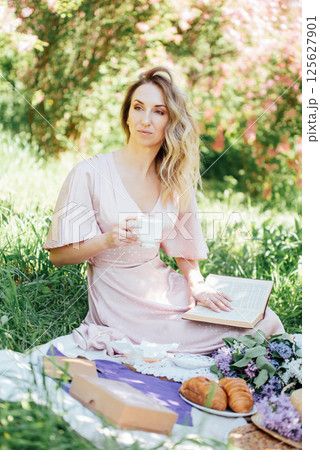 Elegant woman in light pink dress enjoying tea and reading book on spring picnic with croissants, lilac flowers, and gift boxes on grass under blooming trees Elegant woman in light pink dress enjoying tea and reading book on spring picnic with croissants, lilac flowers, and gift boxes on grass under blooming trees 125627901