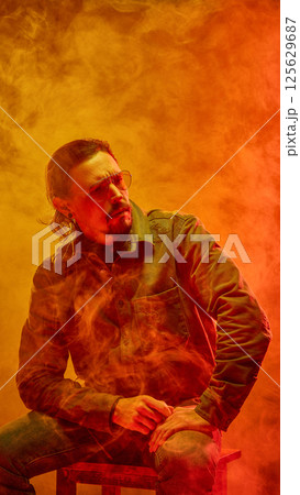Man in glasses sits in smoke lighting in red-orange neon with tense body posture showing resistance and silent anger. 125629687