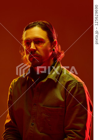 Portrait of young man with distant look in red light with golden glasses shows disconnection from present reality. Portrait of young man with distant look in red light with golden glasses shows disconnection from present reality. 125629690