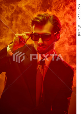 Stylish young businessman in sunglasses lifts shades slowly under thick smoke revealing restrained rage. 125629695