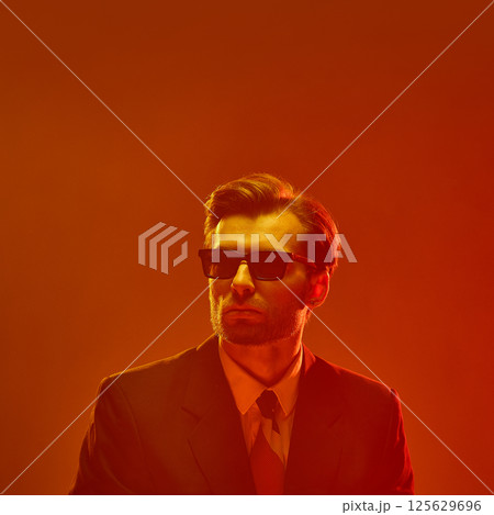 Man in sunglasses with confident chin-up pose against deep orange glow expressing cold detachment Man in sunglasses with confident chin-up pose against deep orange glow expressing cold detachment 125629696