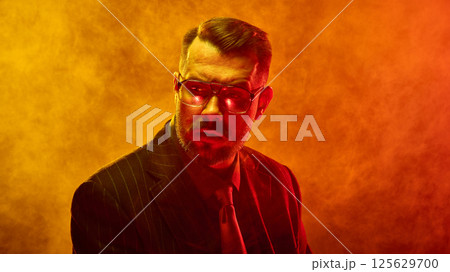 Bearded man in orange-red neon light and fog looks sideways with dramatic tension across face reflecting emotional conflict. Bearded man in orange-red neon light and fog looks sideways with dramatic tension across face reflecting emotional conflict. 125629700