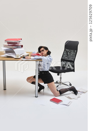 Portrait of young woman looking exhausted resting head on arm under overloaded desk in office environment against white background. Portrait of young woman looking exhausted resting head on arm under overloaded desk in office environment against white background. 125629709