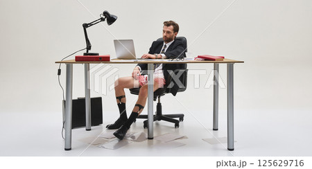 Full length photo of young man in suit jacket and underwear working from home at desk with laptop and serious expression. Full length photo of young man in suit jacket and underwear working from home at desk with laptop and serious expression. 125629716