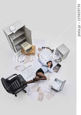 Aerial view photo. Female office worker lying on floor with loose tie and papers scattered around in chaotic workplace scene. 125629730