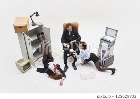 Young woman lying on floor, legs raised inside cabinet while another crawls to boss offering coffee in white surreal office. 125629732