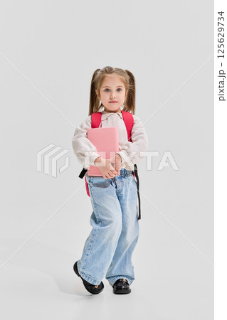 Thoughtful girl with pigtails in sweater holding pink book and walking forward against white background. Thoughtful girl with pigtails in sweater holding pink book and walking forward against white background. 125629734