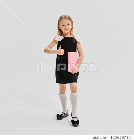 Schoolgirls in stylish outfits with backpacks and books stand showing thumbs up against white background. Concept of guidance Schoolgirls in stylish outfits with backpacks and books stand showing thumbs up against white background. Concept of guidance 125629736