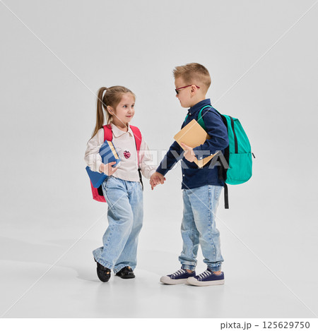 Schoolboy and schoolgirl holding hands with backpacks and books in school outfits walk together against studio background. Schoolboy and schoolgirl holding hands with backpacks and books in school outfits walk together against studio background. 125629750