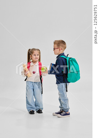 Portrait of little girl holding apple and scroll talks with schoolboy in denim and glasses as they prepare for school lesson against white background. 125629751