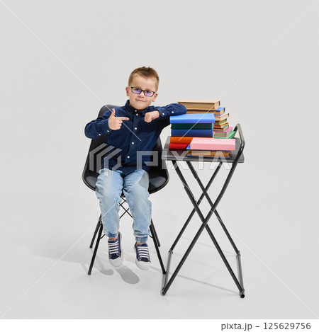 Boy in blue shirt and jeans pointing at colorful books stacked on desk, seated confidently. Book worm. Concept of learning 125629756
