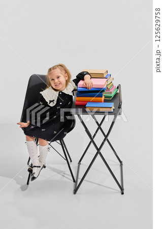 Girl in black dress leans cheerfully on stack of colorful books, smiling confidently at camera. Library. Girl in black dress leans cheerfully on stack of colorful books, smiling confidently at camera. Library. 125629758