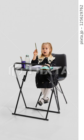 Young girl in black dress sitting at desk raising hand with curious facial expression and open notebooks. Concept of active learning 125629762