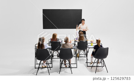 Female teacher explaining in front of blackboard while students listen attentively from desks. Concept of knowledge sharing 125629783