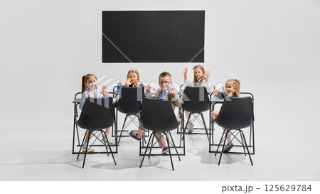 Smiling children sitting at desks facing camera with raised hands in clean classroom setting. Concept of learning, primary education 125629784