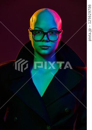 Close-up portrait of of bald woman in stylish dark coat and glasses under vivid studio lights expressing confidence silence strength. 125629786