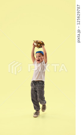 Joyful, triumphant child lifts glove in air after catching baseball during play against pastel yellow background. 125629787