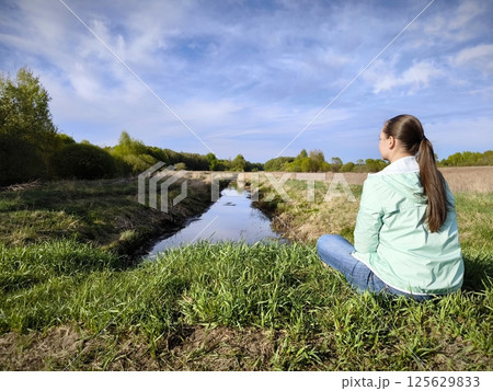young woman is sitting alone in a meadow in early spring, against the background of a calm narrow river. Youth and reflection on life. Girl is sitting alone on the grass by the river. backside view young woman is sitting alone in a meadow in early spring, against the background of a calm narrow river. Youth and reflection on life. Girl is sitting alone on the grass by the river. backside view 125629833