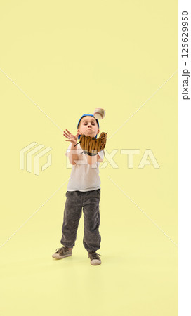 Little boy with glove catching baseball mid-air with focus and excitement against yellow studio background. 125629950