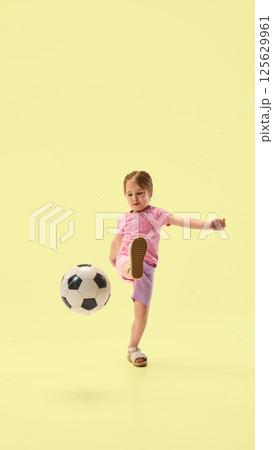 Focused girl in pink raising leg and kicking soccer ball with precision and balance against yellow studio background. Focused girl in pink raising leg and kicking soccer ball with precision and balance against yellow studio background. 125629961