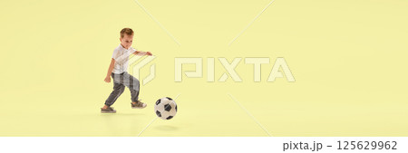 Banner. Athletic young boy kicks soccer ball forward in motion while running across yellow studio background with negative space. 125629962