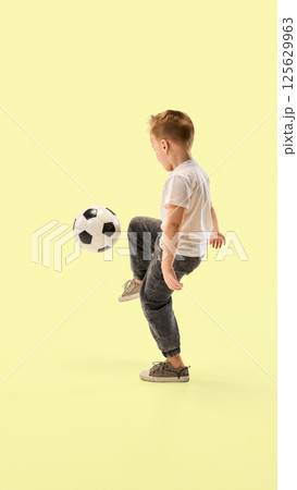 Little champion, sporty boy balances soccer ball with foot focused on action against yellow studio background showing coordination. 125629963