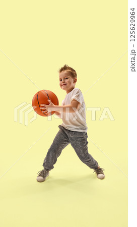 Smiling boy with spiked hair, dressed sportswear, holds basketball ready to play confidently against pastel yellow background. Smiling boy with spiked hair, dressed sportswear, holds basketball ready to play confidently against pastel yellow background. 125629964