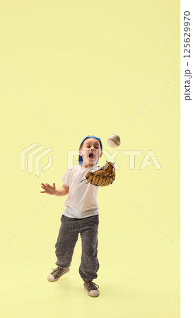 Excited boy, focused on game, dressed in cap catches baseball with glove smiling against yellow studio background. 125629970