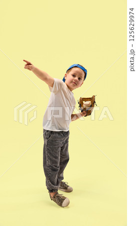 Confident little boy dressed in white shirt pointing direction while holding baseball glove against pastel yellow background. 125629974
