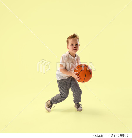 Focused little boy, junior champion with basketball prepares to dribble showing determination under against yellow studio background. 125629997