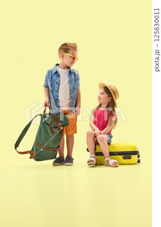 Full length photo of boy standing with backpack and looking at girl sitting on suitcase against pastel yellow background. Full length photo of boy standing with backpack and looking at girl sitting on suitcase against pastel yellow background. 125630011