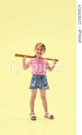 Smiling little girl holding wooden bat behind shoulders standing relaxed and showing confidence against yellow background. Smiling little girl holding wooden bat behind shoulders standing relaxed and showing confidence against yellow background. 125630014