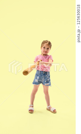 Full length photo of girl in denim shorts confidently swinging wooden bat with smile against pastel yellow background. Full length photo of girl in denim shorts confidently swinging wooden bat with smile against pastel yellow background. 125630018
