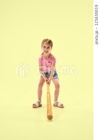 Full length photo of happy girl with pink shirt swinging wooden bat showing joy and energy against pastel yellow background. Full length photo of happy girl with pink shirt swinging wooden bat showing joy and energy against pastel yellow background. 125630019