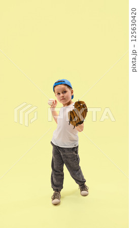 Little boy holding glove and baseball preparing to throw against pastel yellow background. Junior champion training. 125630420