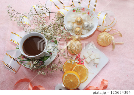 A cup of coffee with lemon and flowers in a festive ribbon, a plate with sweets on a pink background A cup of coffee with lemon and flowers in a festive ribbon, a plate with sweets on a pink background 125634394