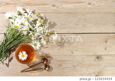 Chamomile tea in a transparent cup with natural small chamomile flowers on a light rustic table, the concept of herbal tea and proper natural nutrition, summer postcard, selective focus 125634434