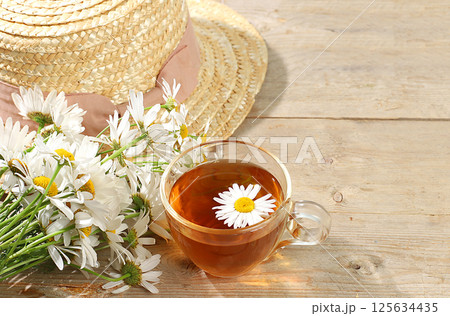 Chamomile tea in a transparent cup with natural small chamomile flowers and a summer hat on a light rustic table, the concept of herbal tea and proper natural nutrition, selective focus 125634435