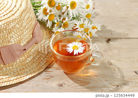 Chamomile tea in a transparent cup with natural small chamomile flowers on a light rustic table, concept of herbal tea and proper natural nutrition, summer card and outdoor recreation, selective focus 125634449