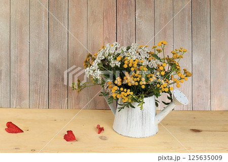 Tansy and yarrow in a watering can on a wooden background, collection of useful herbs for treatment according to folk recipes. Floral arrangement of wild herbs and flowers on a wooden table, rustic 125635009