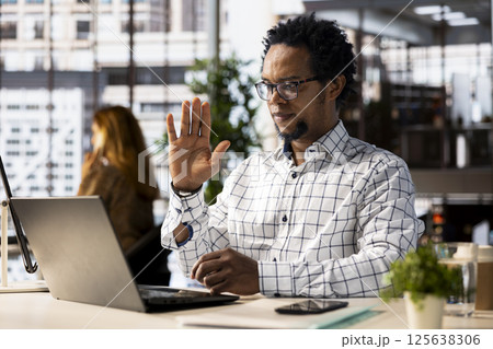 Black man employee joins a telecommuting session at his office desk, multitasking between project management and business strategy discussions. Productivity and collaboration across departments. 125638306