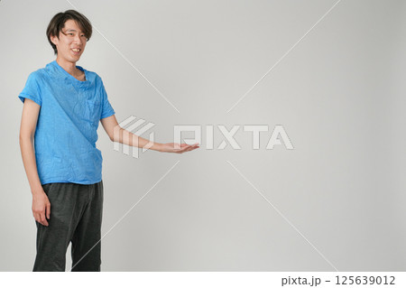 青シャツと黒のジョガーパンツを着た日本人男性がこちらを見て軽く微笑み、左手を腰の高さで上に向けて何か 125639012