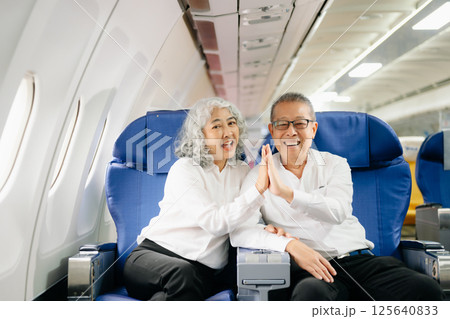 Smiling senior couple of Asian tourists take photos of each other while sitting on a plane. with digital tablet, laptop and smartphone. Travel in style, Smiling senior couple of Asian tourists take photos of each other while sitting on a plane. with digital tablet, laptop and smartphone. Travel in style, 125640833