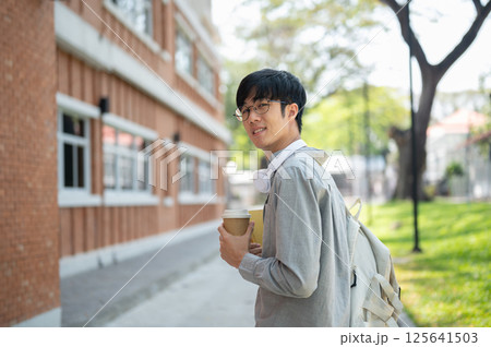 Asian man with glasses is holding a coffee cup and looking at camera while standing near brick wall 125641503