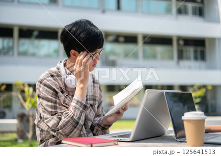 An asian man is touching his glasses while reading a notebook sitting with a laptop at table in park 125641513