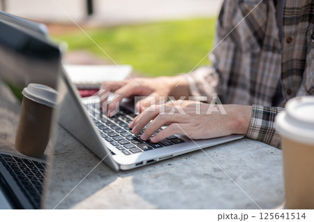 Close up of hands typing on laptop while sitting with friend or coworker at concrete table in park 125641514