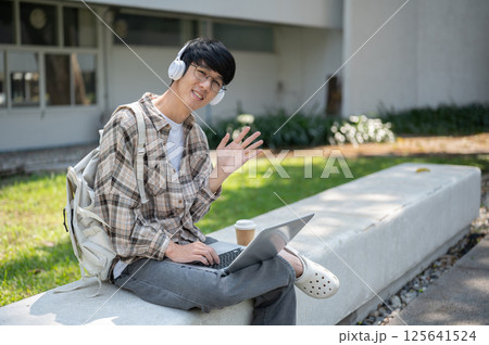 Smiling asian man with glasses and headphone waving hand while sitting with laptop on concrete bench Smiling asian man with glasses and headphone waving hand while sitting with laptop on concrete bench 125641524